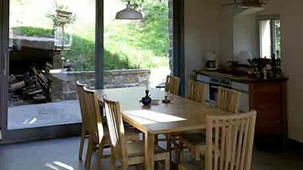 A spacious dining area with a large wooden table and chairs, large windows offering views of the outdoors, and a kitchen area visible in the background.