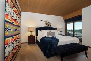 A bedroom with a large bed, bedside lamps, a hanging tribal artifact, and a view through a window.