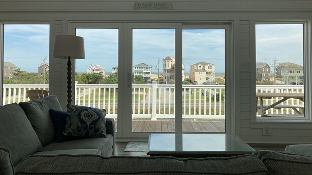 A living room with a large window overlooking a deck and beach houses. - Perfect Ocean and Sunset Views / Deck to Sand in 2 Min (Nags Head)
