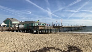 A beach with a wooden pier extending into the ocean, a building with a green roof, and flags on the pier. - Perfect Ocean and Sunset Views / Deck to Sand in 2 Min (Nags Head)