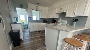 A kitchen with white cabinets, a wooden floor, a microwave, a stove, and a small island with stools. - Perfect Ocean and Sunset Views / Deck to Sand in 2 Min (Nags Head)
