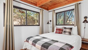 Une chambre avec un grand lit, deux fenêtres, un plafond en bois et une table de chevet avec une lampe.