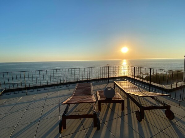 Una terraza con dos mesas de madera y un banco, con vista a un cuerpo de agua al atardecer.