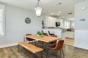 A dining area with a wooden table and benches, a hanging light fixture, and a kitchen in the background.