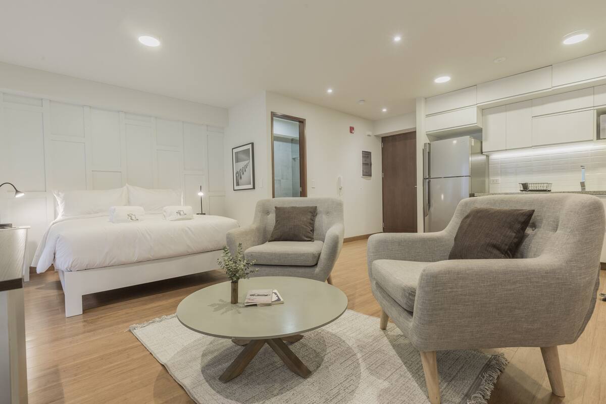 Ein modernes Hotelzimmer mit einem Bett, zwei Sesseln, einem Couchtisch, einer kleinen Pflanze und einer Küchenzeile.