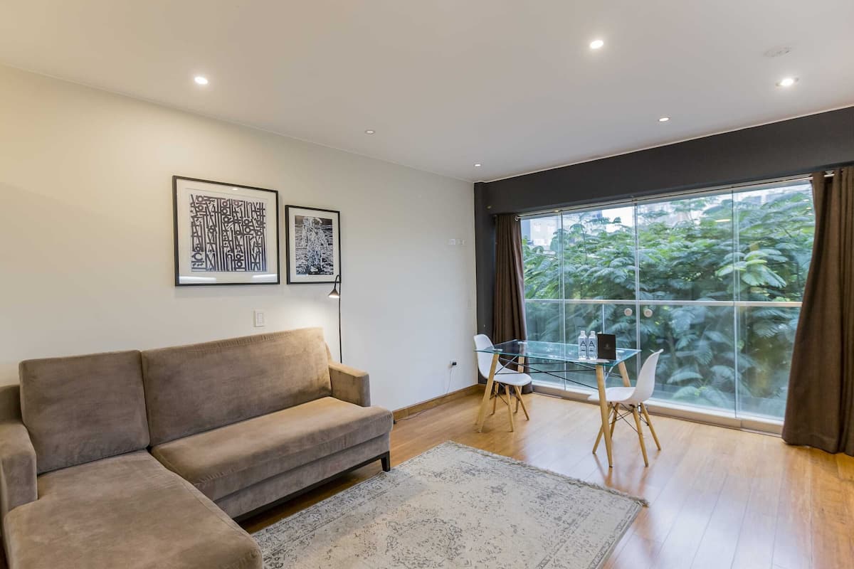 Ein modernes Wohnzimmer mit einer Couch, einem Glastisch, Stühlen, einem großen Fenster mit Vorhängen und zwei gerahmten Kunstwerken an der Wand.