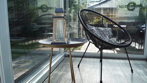 Una habitación moderna con un ventanal que da a un espacio exterior, una pequeña mesa redonda con un farol y una silla negra con cojín.