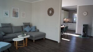 Una sala moderna con un sofá gris, cojines azules, una pequeña mesa redonda y una cocina al fondo.