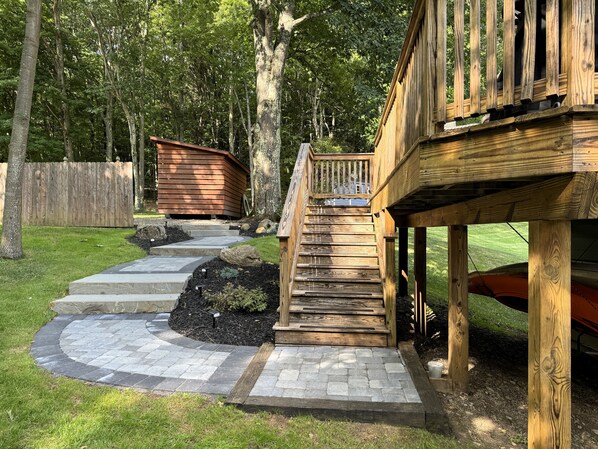 Una terraza de madera con una escalera que lleva a un área cubierta, rodeada por un sendero de piedra y una cerca de madera.