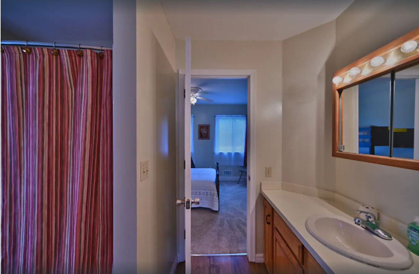Ein Badezimmer mit Duschvorhang, Waschbecken mit Spiegel und einer Tür zu einem Schlafzimmer mit Bett und Fenster.
