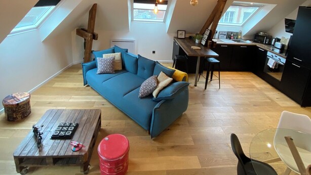 A loft-style apartment with a blue sofa, wooden beams, skylights, and a kitchen area.
