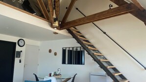 A loft-style room with exposed wooden beams, a staircase, a dining area with a table and chairs, and a blue sofa with patterned cushions.