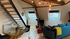 A loft-style living space with a wooden staircase, a glass dining table, a blue sofa, and a television.