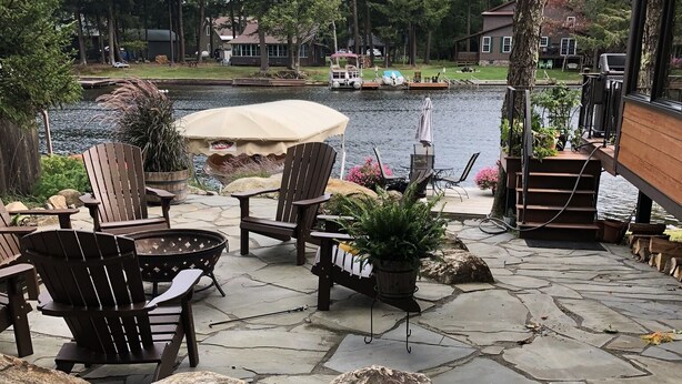 A stone patio with Adirondack chairs and a table, overlooking a lake with boats and houses in the background. - New waterfront house with deck,  dock, close to Old Forge, fireplace, , patio (Old Forge)