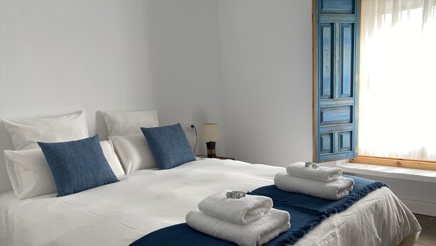 Een net opgemaakt bed met wit- en blauw beddengoed, een blauwe franjes sjaal en twee opgevouwen handdoeken aan het voeteneinde.