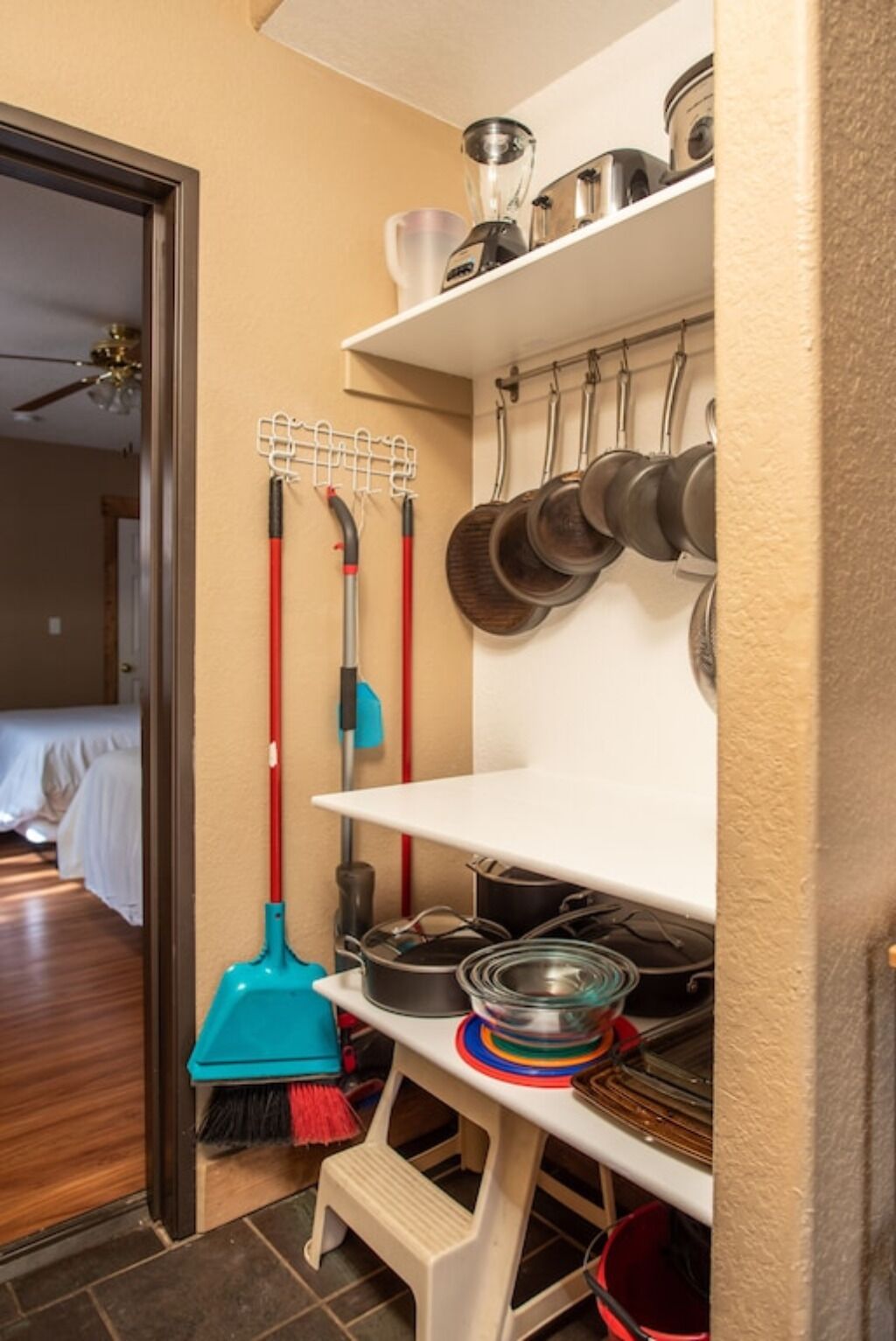 Una cocina con un estante blanco que sostiene ollas y sartenes, un escoba y un mopa.