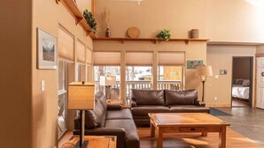 Un salon avec un canapé, une table basse et une lampe. Il y a des fenêtres avec des stores, une étagère avec des objets décoratifs et une porte qui donne sur une autre pièce.