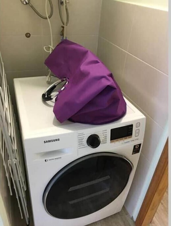 Une machine à laver Samsung dans une salle de bain avec un mur carrelé et un tuyau de douche visible.
