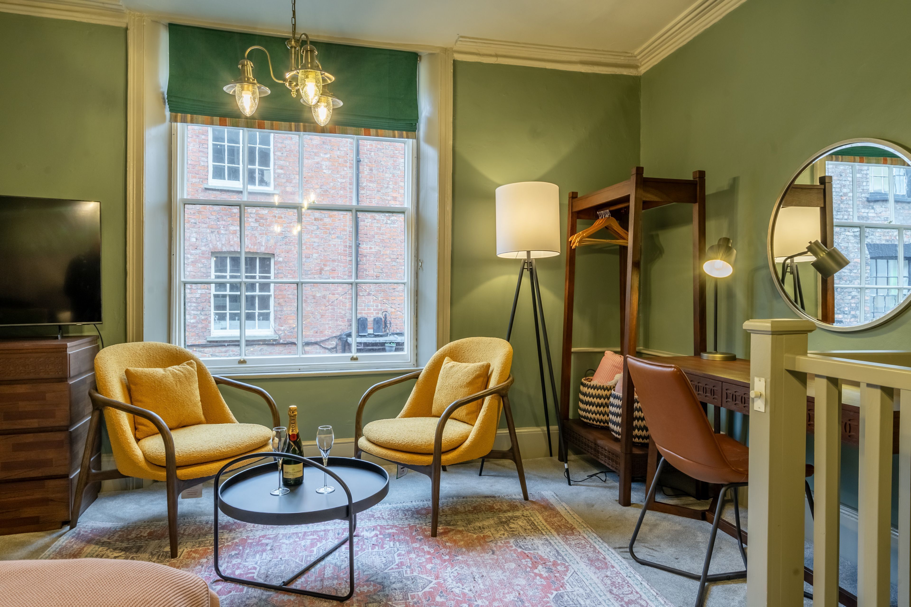 A room with two yellow armchairs, a small round table with a bottle and glass, a floor lamp, a mirror, and a view of a brick building through a window.