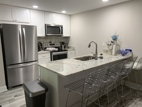 A modern kitchen with stainless steel appliances, a marble countertop, and a bar area with metal chairs.