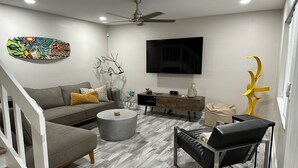 A modern living room with a grey sofa, a round coffee table, a black ottoman, a wooden cabinet, and a flat-screen TV mounted on the wall.
