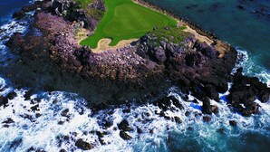 Vista aérea de un campo de golf costero con una isla rocosa en primer plano y un cielo azul despejado.