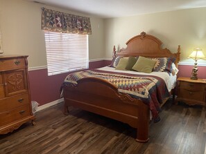 A bedroom with a wooden bed, a nightstand, a dresser, a window with blinds, and a quilted headboard.