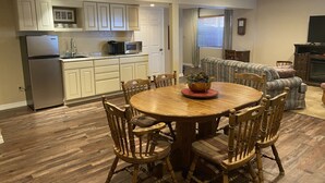 A compact kitchen with a refrigerator, microwave, and sink. There is a dining table with chairs and a potted plant. A sofa and a television are visible in the background. - Spacious and Comfortable Garden Level Basement Apartment (Peyton)