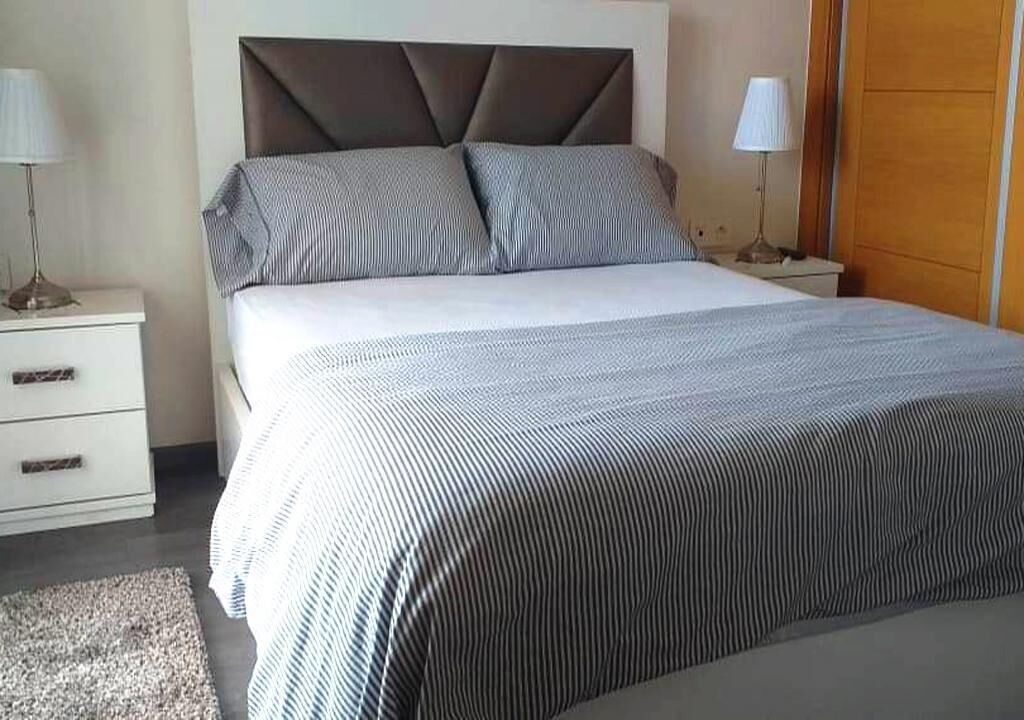 A neatly made bed with a striped bedspread and two pillows, a bedside table with a lamp, and a wooden wardrobe in a hotel room.
