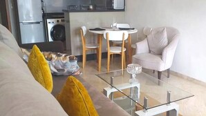 A living room with a glass coffee table, a sofa with yellow cushions, a white armchair, and a dining area with a white table and chairs. - Residence Hivernage Founty 4773 (Agadir)