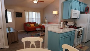 Una sala de estar con un sofá rojo, una mesa de centro y una zona de comedor con una mesa y sillas blancas. La cocina tiene armarios azules y electrodomésticos blancos.