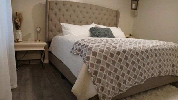 Un dormitorio con cabecera capitonada, cama con ropa de cama estampada, mesita de noche con lámpara y un cartel en la pared.