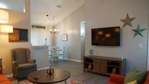 A living room with a sofa, armchair, coffee table, and a TV mounted on the wall. There is a chandelier, a sign that reads "FARMHOUSE," and a star decoration on the wall.