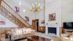Un salon spacieux avec un escalier en bois, une cheminée, un téléviseur et un lustre.
