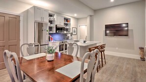 A modern kitchen with white cabinets, a stainless steel refrigerator, and a microwave. There is a dining table with chairs and a TV mounted on the wall.
