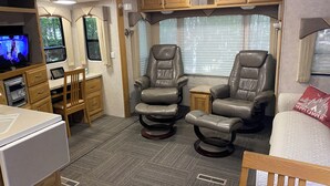 Un intérieur de camping-car compact comprenant un coin cuisine, deux fauteuils inclinables, une petite table et un lit agrémenté d’un coussin orné de la mention « Happy Camper ».