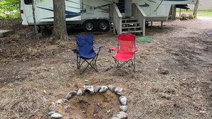 Un camping-car blanc avec un porche couvert, deux chaises pliantes et un foyer au milieu d’une zone boisée.