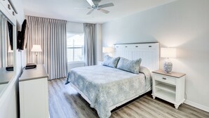 Une chambre Ă coucher avec un lit, des tables de chevet, un ventilateur de plafond et une fenĂȘtre avec des rideaux.