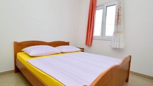 Una cama individual con estructura de madera y colchón amarillo, ropa de cama blanca y una ventana con cortinas naranjas.