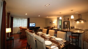 A dining area with a long table set for a meal, a television in the background, and a kitchen area with a stove and sink. - Kingfisher Cottage - Russell Cottages Collection (Russell)