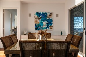 A dining area with wicker chairs, a glass table, and a blue abstract painting on the wall. - Sunshine Coast White House (Castaways Beach)