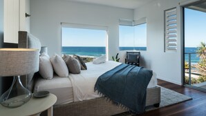A bedroom with a large bed, a nightstand, a lamp, and a view of the ocean through large windows. - Sunshine Coast White House (Castaways Beach)