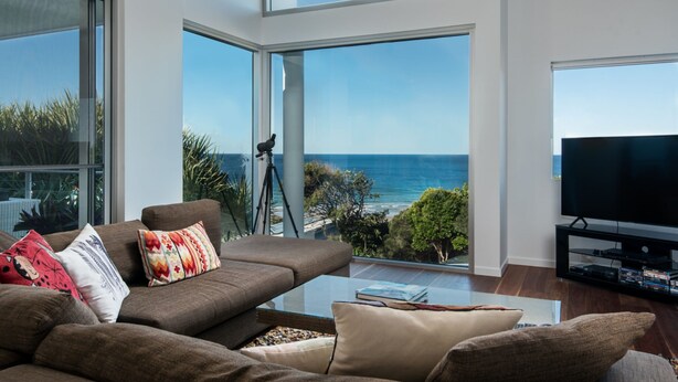A modern living room with a large window overlooking the ocean, a comfortable sofa, and a flat-screen TV on a stand. - Sunshine Coast White House (Castaways Beach)