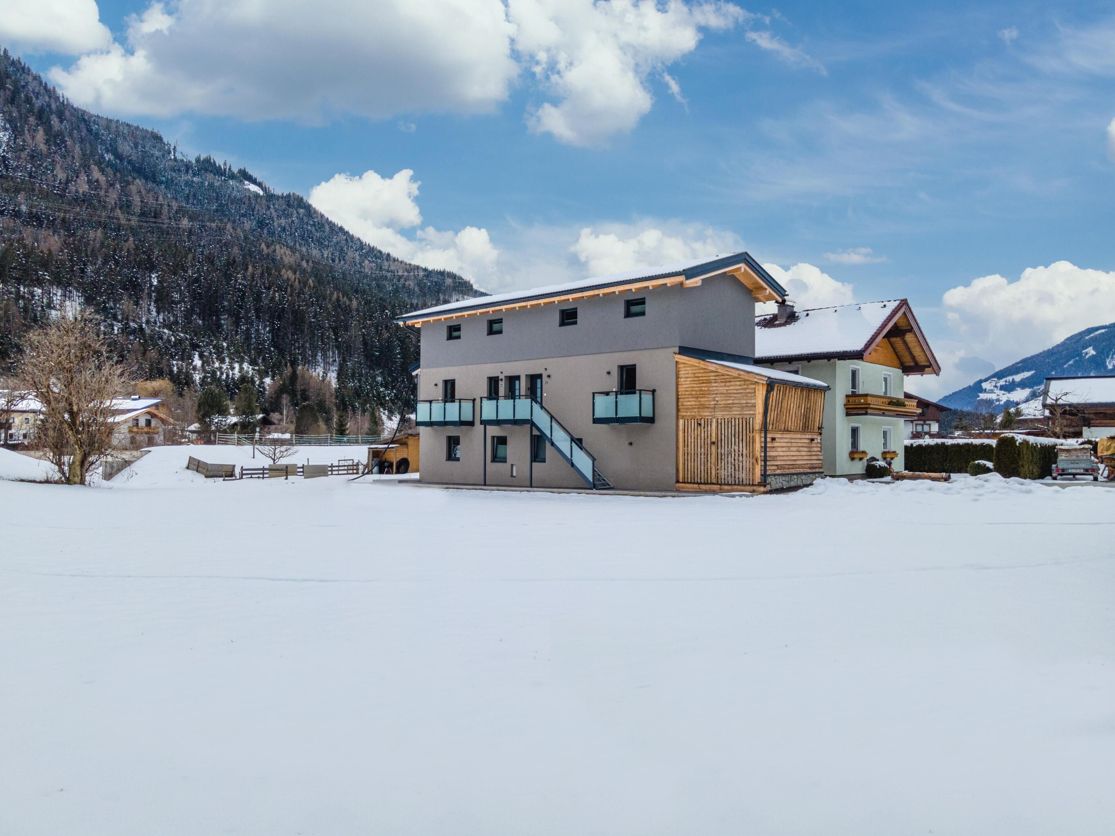 Un edificio de dos pisos con un balcón y fachada de madera, rodeado de nieve y montañas al fondo.