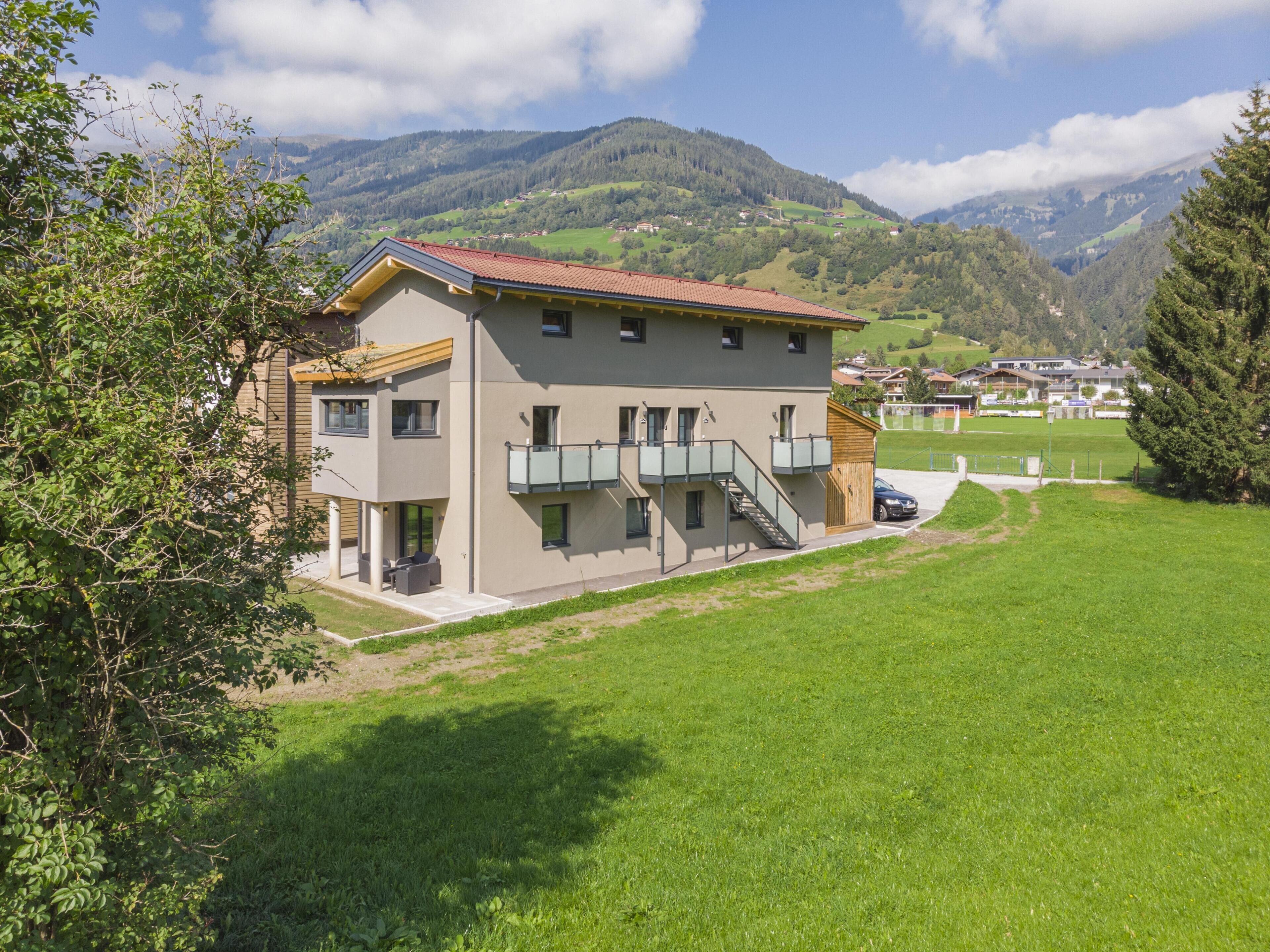 A two-story building with a balcony and a carport, surrounded by green fields and mountains in the background.