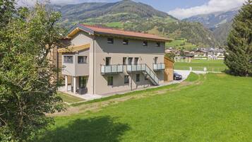 A two-story building with a balcony and a carport, surrounded by green fields and mountains in the background.