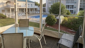 Un espacio al aire libre cubierto con una mesa y sillas de vidrio, un ventilador de techo y vista a una piscina y edificios al fondo.
