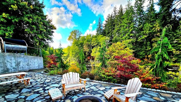 A stone patio with Adirondack chairs and a fire pit, surrounded by lush greenery and trees. - The Cullen's Clair de Lune, River-front Estate (Forks)