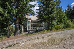 A lodge with a large deck, surrounded by tall trees and a gravel path.
