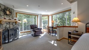 Amplia habitación con chimenea, un sillón de cuero, un escritorio con lámpara y ventanas grandes que dan a vistas de árboles.